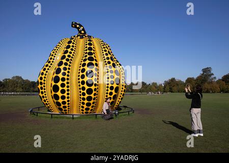 Eine Person fotografiert eine andere vor Yayoi Kusamas Kürbis am Round Pond in Kensington Gardens, London. Die Skulptur ist gelb und schwarz. Stockfoto