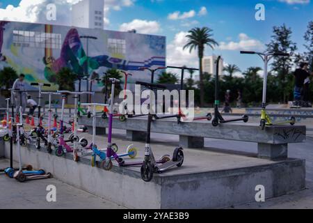 Eine Sammlung von farbenfrohen Motorrollern, die auf Betontreppen in einem städtischen Außenbereich angeordnet sind, mit einem lebhaften Wandgemälde und Palmen im Hintergrund. Stockfoto