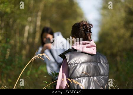 Mom macht ein Foto von einem kleinen Mädchen in Sweatshirt und Weste, das mitten auf einem Weg in einem dichten Wald steht. Das Konzept einer fröhlichen Familie überhaupt Stockfoto