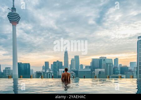 Rückansicht von Mann reist und entspannt sich im Infinity-Pool auf dem Dach bei Sonnenaufgang, Kuala Lumpur, Malaysia. Männlicher Reisender, der die asiatische Stadtlandschaft genießt Stockfoto