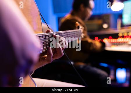 Ein talentierter Künstler spielt neue Akkorde auf seiner Elektro-Akustikgitarre im Studio und nimmt neue Tracks mit dem Musikproduzenten im Kontrollraum auf. Männlicher Künstler, der Melodien auf dem Instrument komponiert. Nahaufnahme. Stockfoto