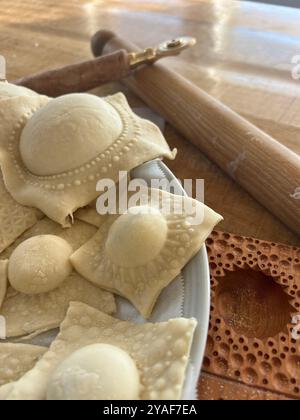 Frisch zubereitete Ravioli, die auf einem Teller neben einem Holzrollstift und einem Ravioli-Cutter liegen, bereit für den nächsten Schritt in der Nudelherstellung Stockfoto