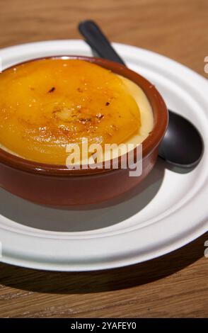 Katalanische Creme serviert in einer Tonschale mit einem schwarzen Löffel auf hölzernem Hintergrund. Selektiver Fokus Stockfoto