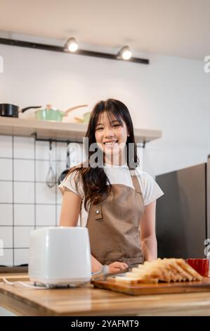 Ein Porträt einer schönen jungen asiatischen Frau, die ihr Frühstück in der Küche zubereitete, Toast und Kaffee zubereitete und in die Kamera lächelte. Zuhause leben, Kuppeln Stockfoto