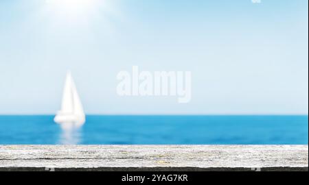 Ein leerer Holztisch im Vordergrund mit einem sonnigen Meereshorizont und einem Segelboot im Hintergrund. Der klare blaue Himmel und das ruhige Meer bieten Ihnen etwas Stockfoto