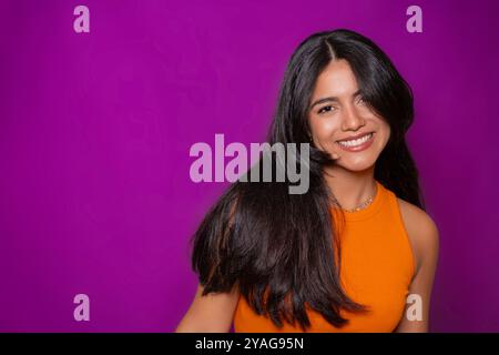 Fröhliche Frau mit fließenden Haaren, lässiges orangefarbenes Oberteil, warm lächelnd, lila Hintergrund Stockfoto