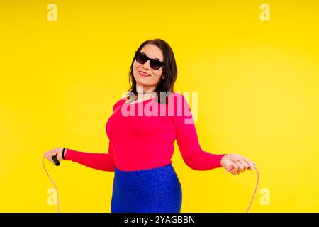 Hübsch lächelnde, pralle Frau in sportlichem Oberteil und Leggings, die ein Springseil in den Händen hält Stockfoto