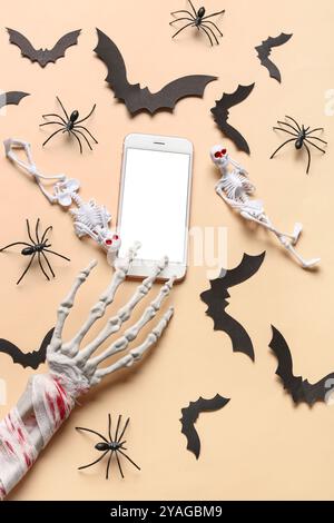 Komposition mit Skeletten, Hand, modernem Mobiltelefon, Fledermäusen und Spinnen für die Halloween-Feier auf beigefarbenem Hintergrund Stockfoto