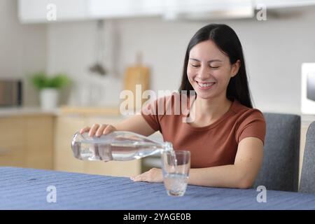 Glückliche asiatische Frau, die ein Glas Wasser aus der Flasche in der Küche zu Hause füllt Stockfoto