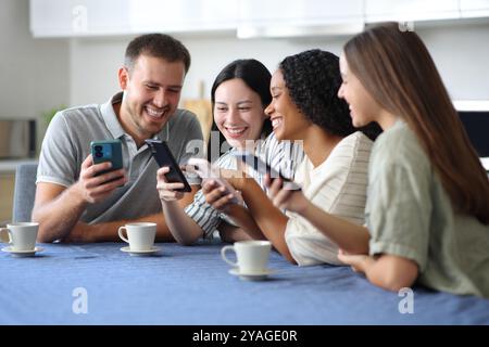 Vier fröhliche Freunde, die in der Küche stehen und zu Hause Telefone überprüfen Stockfoto