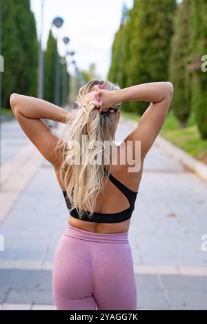 Die blonde Frau bindet sich die Haare, während sie sich auf ihre Trainingsroutine im Park vorbereitet Stockfoto