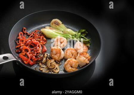 Gebratene Garnelen und Gemüse wie Pak Choy, rote Paprika und Zwiebeln in einer Pfanne auf einem schwarzen Induktionsherd, kochen eine gesunde Mahlzeit für niedrige C Stockfoto