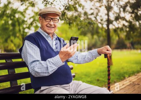 Porträt eines glücklichen älteren Mannes im Park. Er sitzt auf der Bank und benutzt ein Smartphone. Stockfoto