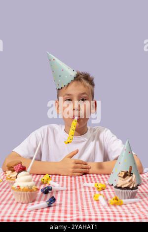 Ein süßer Junge mit Partyhut bläst ein Horn auf einem Geburtstagskuchen am Tisch. Glückliches Teenager-Kind Stockfoto