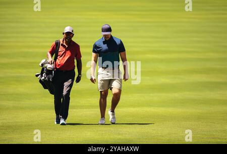 Ein Caddy und ein Golfer gehen Seite an Seite und diskutieren auf einem hellen Golfplatz nachmittags bei einer Wettkampfrunde bei strahlendem Sonnenschein über Strategie. Stockfoto