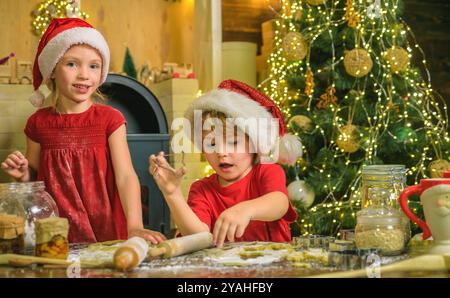 Lustige Kinderköche in weihnachtsmützen bereiten das Weihnachtsessen zu. Zwei Kildren in weihnachtsmänteln und Schürzen bereiten gemeinsam weihnachtskekse vor. Stockfoto