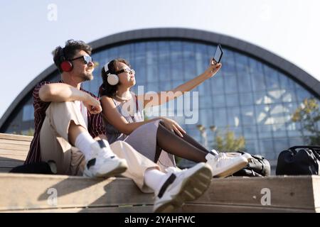 Ein junges Paar mit Kopfhörern hört Musik und macht ein Selfie im Freien. Sie sitzen auf Holztreppen mit moderner Architektur im Hinterhaus Stockfoto