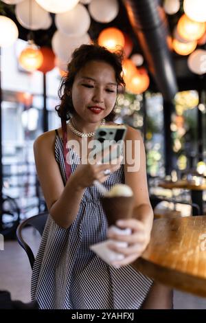 Eine junge Frau mit einem Smartphone lächelt, während sie in einem trendigen Café Eis hält, umgeben von bunten Lichtern und einem lebhaften Ambiente. Stockfoto