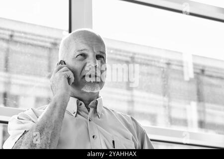 Selbstbewusster, reifer Geschäftsmann, der in modernen Büros telefoniert und Führungsstärke und Erfahrung zeigt. Das Schwarzweißbild strahlt Professionalität und Su aus Stockfoto