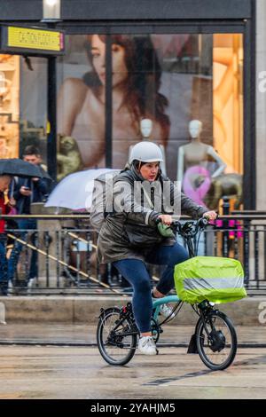 London, Großbritannien. Oktober 2024. Ein elektrisches Brompton-Fahrrad fährt an frühen Käufern vorbei, die trotz des starken Regens auf der Oxford Street unterwegs sind - das feuchte Herbstwetter trifft London weiterhin. Anoraks, Hoodies, Regenschirme, Ponchos sind an der Tagesordnung. Guy Bell/Alamy Live News Stockfoto