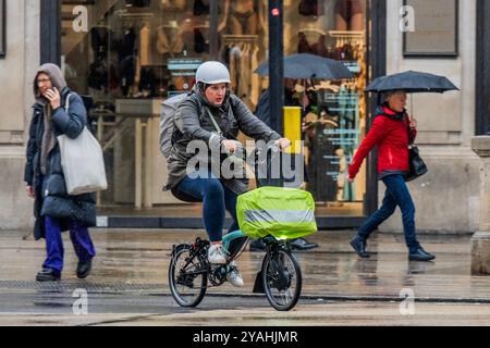 London, Großbritannien. Oktober 2024. Ein elektrisches Brompton-Fahrrad fährt an frühen Käufern vorbei, die trotz des starken Regens auf der Oxford Street unterwegs sind - das feuchte Herbstwetter trifft London weiterhin. Anoraks, Hoodies, Regenschirme, Ponchos sind an der Tagesordnung. Guy Bell/Alamy Live News Stockfoto