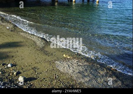 Das Bild zeigt eine Welle, die sanft auf einen Kiesstrand schlägt. Das Wasser ist klar und blau und der Strand ist sandig mit kleinen Felsen, die verstreut sind. Stockfoto