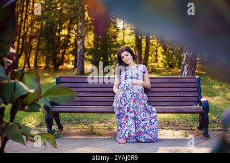 Portret von schwangeren Frauen im Kleid sitzt auf der Bank im warmen Park Stockfoto