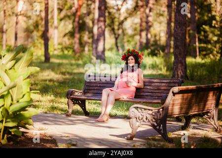 Portret von schwangeren Frauen im Kleid mit Kranz im Kopf sitzt auf einer Bank im warmen Park Stockfoto