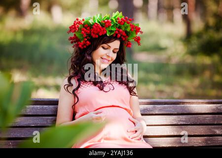 Portret von schwangeren Frauen im Kleid mit Kranz im Kopf sitzt auf einer Bank im warmen Park Stockfoto