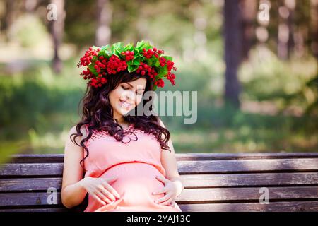 Portret von schwangeren Frauen im Kleid mit Kranz im Kopf sitzt auf einer Bank im warmen Park Stockfoto