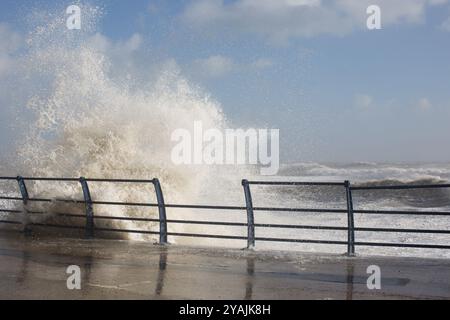 Eine mächtige Meereswelle stürzt gegen das Metallgeländer einer Strandpromenade und schickt an einem stürmischen Tag an der Küste Wasserstrahl hoch in die Luft. Stockfoto