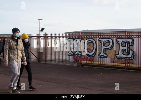 Ein Paar, das Hand in Hand geht mit Gesichtsmasken, das Wort HOFFNUNG im Hintergrund auf den Hastings Pier Geländern. Stockfoto