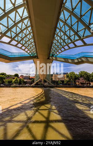 Blick auf das Glasdach, den Fußgängerweg und die Treppen der berühmten Friedensbrücke und die Schatten auf dem Fluss Kura tiflis georgien Stockfoto