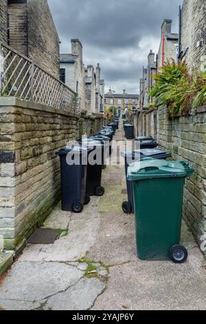 Wheelie-Mülltonnen säumen den Ginnel (Galleyway, snicket) zwischen zwei Reihen von Terrassenhäusern (ehemalige Salzmühle Workers Housing) in Saltaire, Yorkshire. Stockfoto