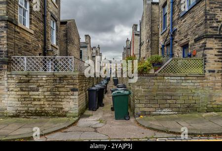 Wheelie-Mülltonnen säumen den Ginnel (Galleyway, snicket) zwischen zwei Reihen von Terrassenhäusern (ehemalige Salzmühle Workers Housing) in Saltaire, Yorkshire. Stockfoto