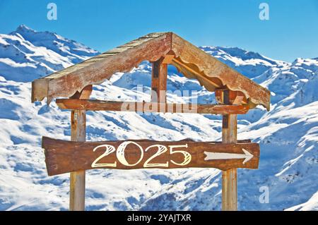 2025 geschrieben auf einem hölzernen Richtungsschild, schneebedeckte Berglandschaft im Hintergrund, Skiferien Neujahrskarte Stockfoto