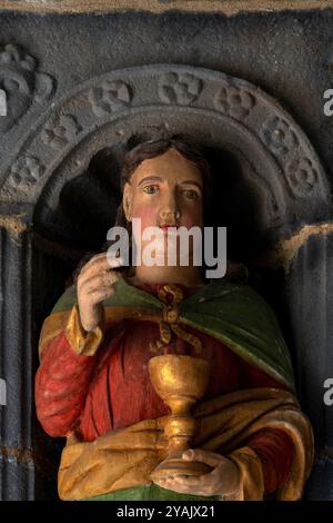 Der Heilige Johannes der Apostel und der Evangelist mit einem goldenen Becher oder Kelch. Kopf und Oberkörper einer von 12 polychromen Apostelstatuen, die 1721 in Holz gemeißelt wurden und den Eingang einer bretonischen Kirche, L’Eglise Saint Sauveur, in Le Faou, Finistère, Bretagne, Frankreich, flankieren. Die Statuen wurden kürzlich restauriert und neu gestrichen. Stockfoto