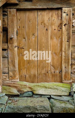 Eine alte Holztür mit horizontalen Dielen und einem Metallschloss ist in eine Holzwand eingefasst. Die Tür ist leicht offen und zeigt einen Steinweg. Stockfoto
