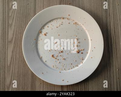 Runde weiße Platte mit Brotkrümeln auf braunem Holztisch, Reste-Konzept, Hungry Games Perspektive Stockfoto