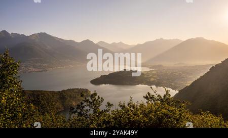 An einem frühen Sommerabend untergeht die Sonne sanft über dem Comer See und strahlt warme Farbtöne über das ruhige Wasser und die umliegenden Berge aus, was eine friedliche Atmosphäre zum Entspannen schafft. Stockfoto