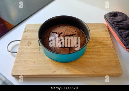 Ein frisch gebackener Schokoladenkuchen, der in einer runden blauen Backform zerbrochen ist, auf einem Holzschneidebrett neben einem Induktionsherd und einem schwarzen Ofenhandschuh platziert wird, Stockfoto