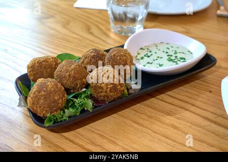Knusprige Falafelbällchen serviert auf einer schwarzen Platte mit grünem Salat und einer Gurke, Joghurt und Minzsauce mit gehacktem Schnittlauchschnitt auf einem Holztisch in A re Stockfoto