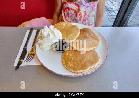 Weißer Teller mit drei Pfannkuchen, Karamell und Schlagsahne auf einem Tisch mit Messer und Gabel, bereit zum Essen von einem kleinen Mädchen Stockfoto