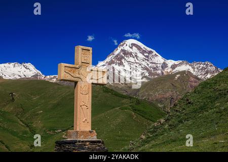 Ein großes gemeißeltes Steinkreuz thront auf einem Hügel über dem Dorf Stiefantsminda mit dem schneebedeckten Berg kazbegi im Hintergrund Stockfoto