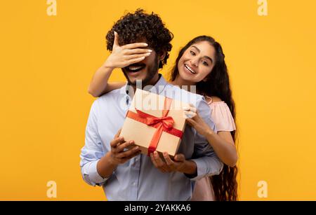 Hübsche indische Frau bedeckt die Augen ihres Freundes, hält eine Geschenkbox und begrüßt ihn mit Geburtstag oder Jahrestag Stockfoto