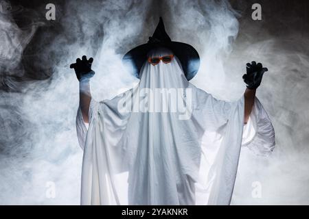 Mann im gruseligen Geisterkostüm für Halloween und Hexenhut auf dunklem mystischen Hintergrund in Rauch, Konzept von Halloween Stockfoto