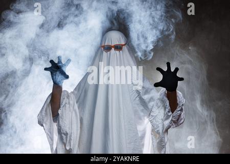 Mann im gruseligen Geisterkostüm in Rauch auf grauem mysteriösem Hintergrund im Studio. Minimales halloween-Geisterkonzept, Konzept von Halloween Stockfoto