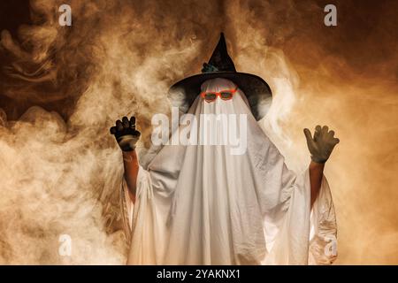 Mann im gruseligen Geisterkostüm für Halloween und Hexenhut auf orangem mystischen Hintergrund in Rauch, Konzept von Halloween Stockfoto