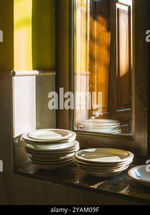 Stapel weißer Teller auf einer Granittheke, in warmes Sonnenlicht getaucht, das durch ein Holzfenster filtert. Stockfoto