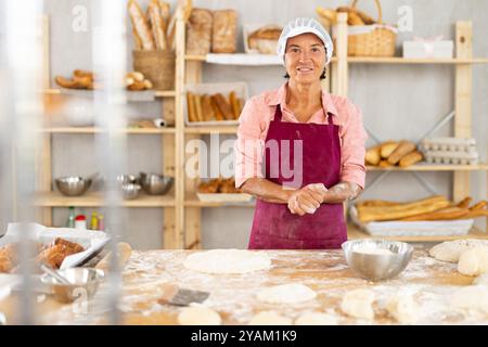 Lächelnde alte Bäckerin, die am bemehlten Tisch in der Bäckerei steht Stockfoto